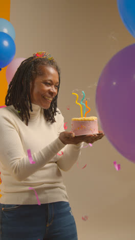 Vertical-Video-Studio-Shot-Of-Woman-Wearing-Birthday-Headband-Celebrating-Birthday-Blowing-Out-Candles-On-Cake-With-Paper-Confetti